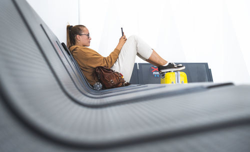 Woman using mobile phone while sitting on bench in airport