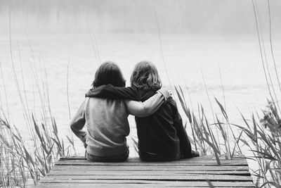 Rear view of brothers sitting with arms around on jetty in lake