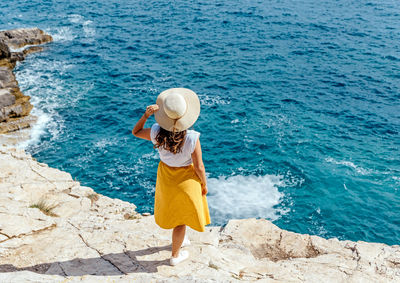 Young woman in summer clothes and hat standing on edge of cliff above sea.