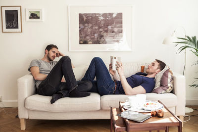 Young gay man reading book while partner looking at him on sofa