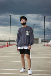 Portrait of young man wearing face mask standing on road against cloudy sky