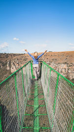 Rear view of woman with arms outstretched on footbridge against blue sky