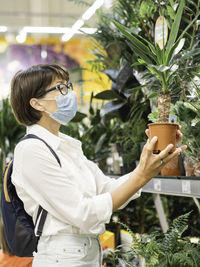 Woman in medical protective mask chooses plants for home. shelves with seedlings, flowering plants. 
