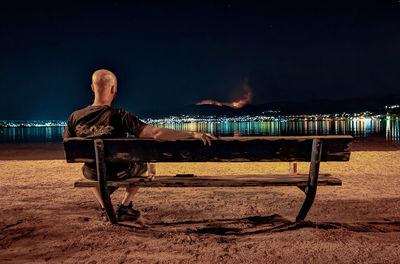 Man sitting on bench at beach against sky at night