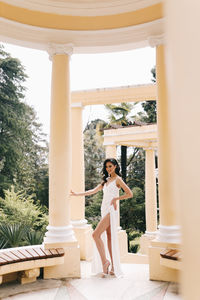 A beautiful brunette lady in an elegant wedding dress poses among the columns in the old city park