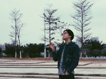 Young man looking away while standing on tree against sky