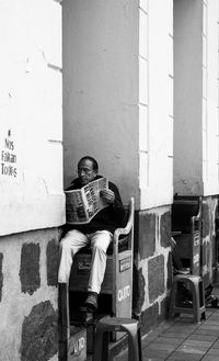 Man sitting on seat reading newspaper against wall