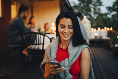 Smiling mid adult woman using mobile phone while friends in background during dinner party