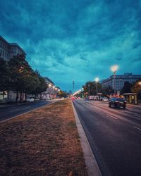 Road by illuminated buildings against sky at dusk