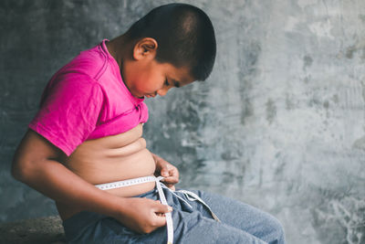 Side view of boy measuring stomach while sitting against wall