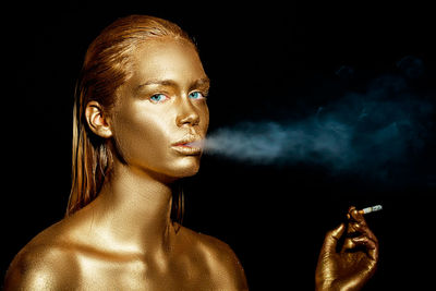 Close-up portrait of woman with golden glitter make-up smoking against black background