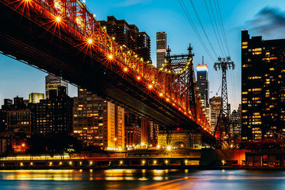 Illuminated bridge over river by buildings against sky at night