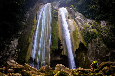 Side view of man sitting on rocks by waterfall in forest