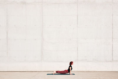 Side view of trendy fit female athlete in sportswear doing baby cobra yoga exercise on sports mat while training alone on street against concrete wall in sunny day