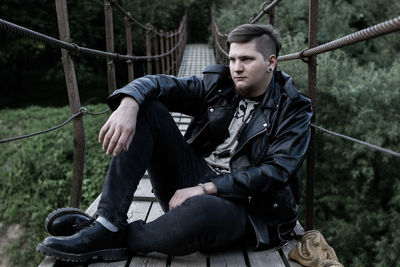 Young man looking away while sitting on railing against trees
