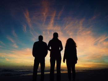 Rear view of silhouette friends standing at beach during sunset