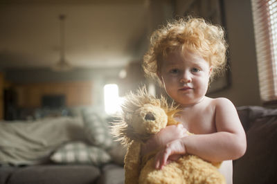 Portrait of cute shirtless baby boy holding stuffed toy in living room at home