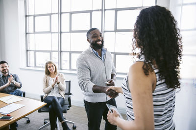 Businessman and businesswoman shaking hands while coworkers applauding in board room