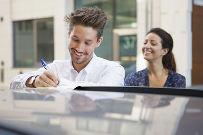 Businessman writing in book on car while female colleague looking at him