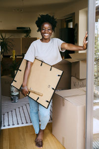 Portrait of happy woman holding frame while leaning on wall at new home