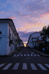 Road by buildings against sky during sunset