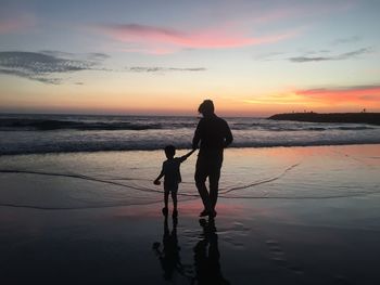 Silhouette father and son walking on beach against sky during sunset