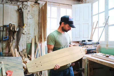 Bearded male artisan in cap with plank for longboard standing against assorted tools and windows in workshop