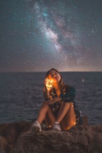 Portrait of young woman sitting on rock at beach against sky at night