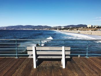 Empty bench on pier by sea against clear blue sky