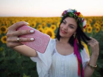 Smiling woman wearing flowers taking selfie through mobile phone while standing on field