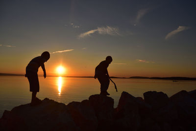 Silhouette of boys standing on rock by sea against sky during sunset