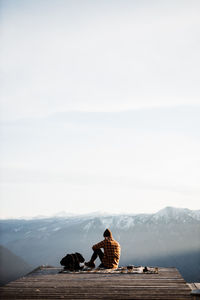 Back view of unrecognizable male tourist sitting on blanket at viewpoint and enjoying scenery of mountain ridge in morning