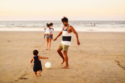 Full body of positive latin american father kicking ball while playing with little son on sandy beach near waving sea