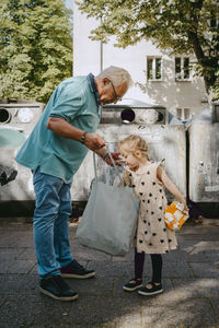Grandfather and granddaughter searching in tote bag while standing in front of garbage bins at street