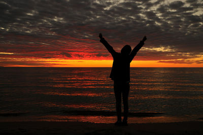 Happy woman sihouette with arms raised up in success to sunset glow at beach by water