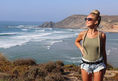 Young woman wearing sunglasses standing at beach against sky