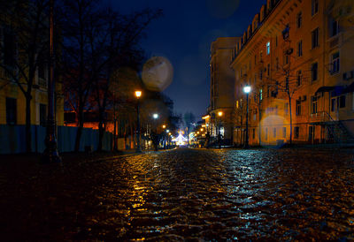 Illuminated street amidst buildings against sky at night