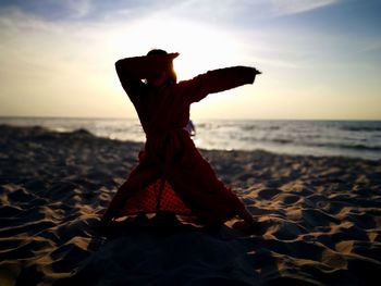 Girl with arms raised exercising at beach against sky during sunset