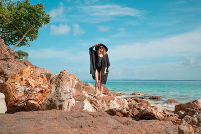 Full length of woman standing on rock at beach against sky