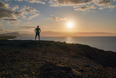 Rear view of man walking at beach against sky during sunset