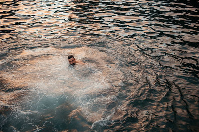 From above of ethnic male with dark hair and beard swimming in dark rippling water and looking at camera in azores