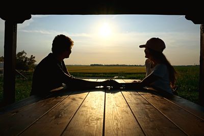 Side view of young couple sitting on landscape against sunset sky