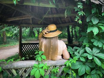 Young woman with long blonde hair and straw hat standing in a gazebo surrounded by green leaves