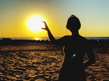 Optical illusion of silhouette woman holding sun while standing at beach against sky during sunset