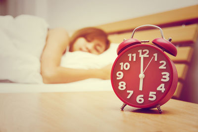 Close-up of alarm clock on table with woman sleeping on bed at home