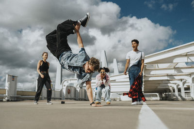 Acrobatic young man doing handstand while male and female friends cheering in parking lot under cloudy sky