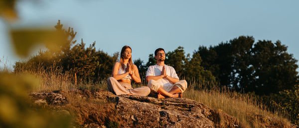 Man and woman meditating outdoors while sitting on rock, adult couple practicing yoga at sunset	
