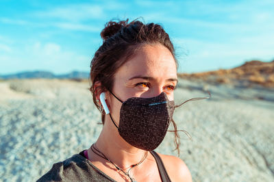 Side view of confident young sportswoman in black protective mask listening to music with wireless earbuds while recreating during training in rough desert badlands looking away