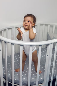 One year old child in white clothes standing in a white round bed in his nursery