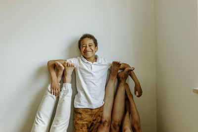 Happy boy with hands on parent's and sister's legs in front of wall